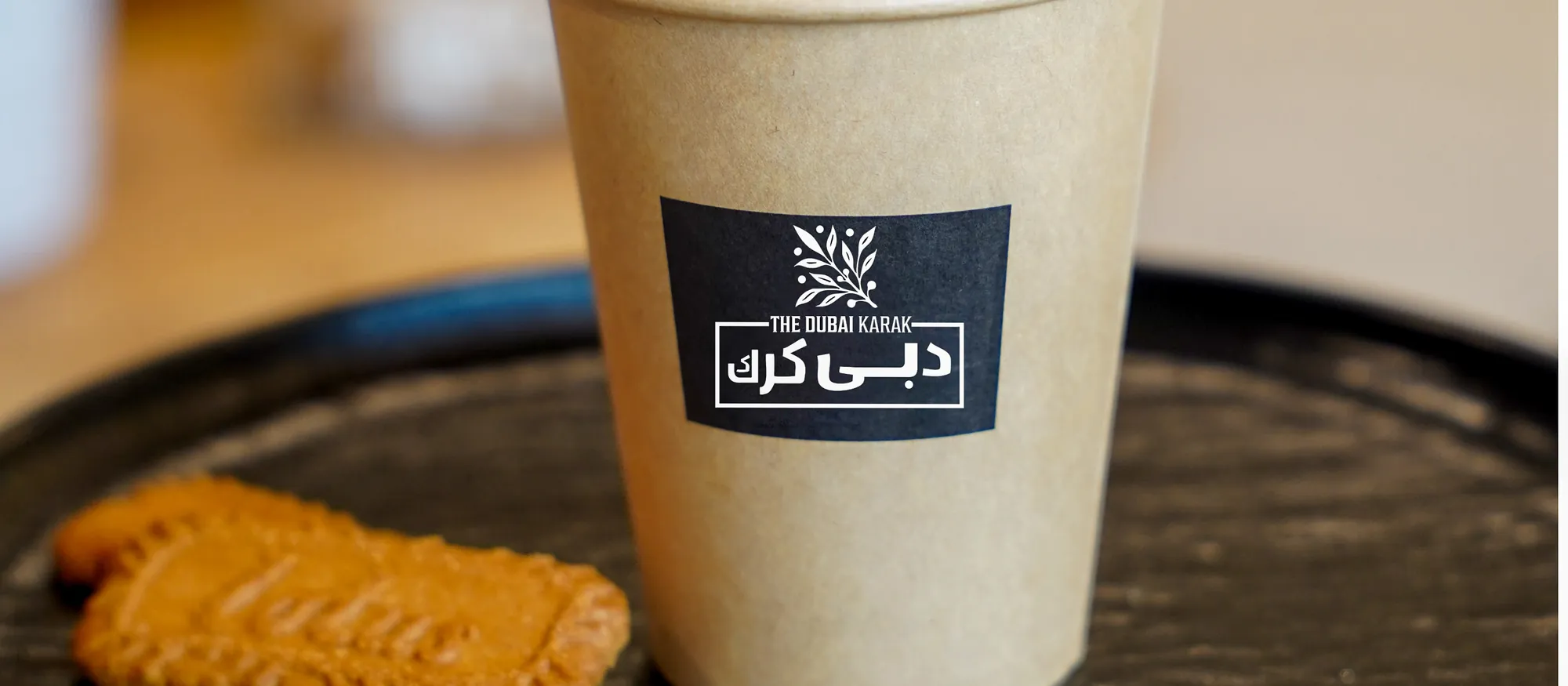 A branded cup of Dubai karak chai served on a black tray with a Lotus biscuit