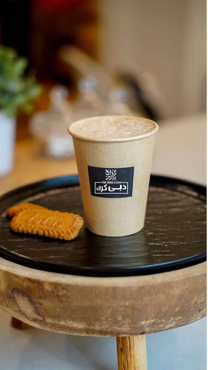 A branded cup of Dubai karak chai served on a black tray with a Lotus biscuit